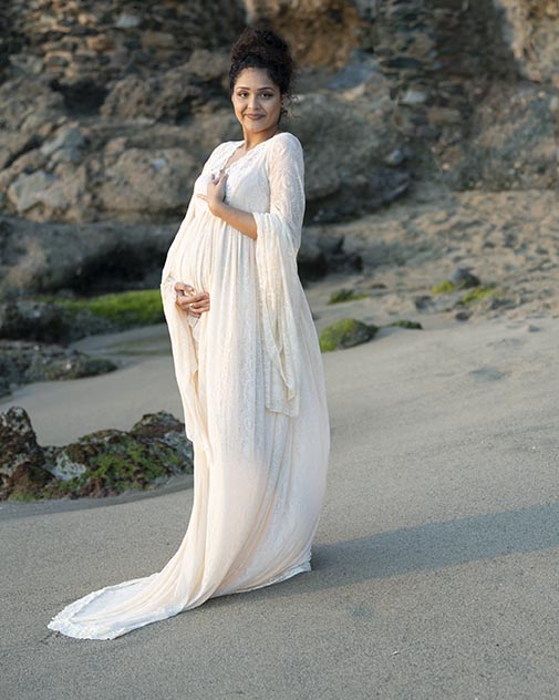 A pregnant woman in a long white dress isstanding on the beach in the Bay area.