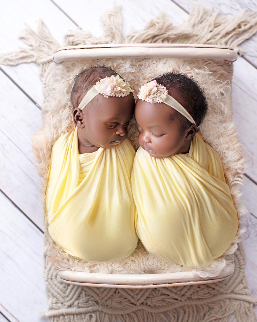 Twin newborn girls wrapped in a yellow wrap and laying on a small bed.