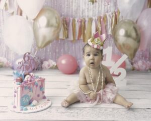 A baby girl is sitting next to a birthday cake with balloons in the background.