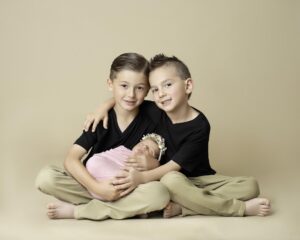 Boys wearing khaki pants and black tee shirts sitting on a floor holding their newborn baby sister in their laps after visiting Orange County Baby Stores.