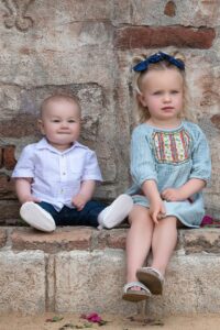 A happy baby in a white shirt sits on a stone ledge with his older sister in a blue dress