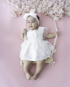 A happy baby smiles while sitting on a swing in a white lace dress and matching bow after visiting Orange County Baby Stores