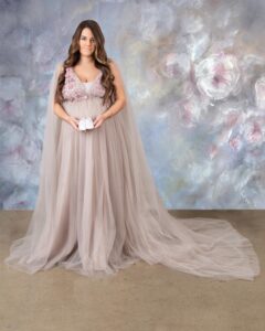 A pregnant woman in the Bay Area is in a beige gown is olding a pair of baby shoes.