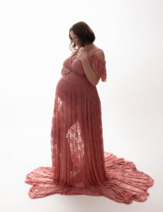 A pregnant woman in the East Bay area is wearing a rose colored lacy maternity gown