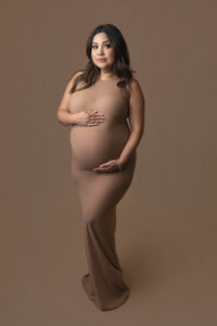 A pregnant woman in the Bay area is wearing a beige gown and standing.