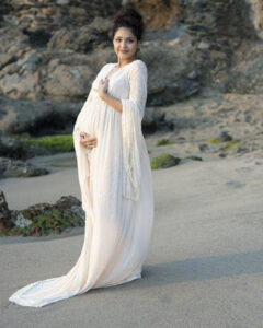 A pregnant woman in the Bay area is wearing a long white gown. She is standing on a beach.