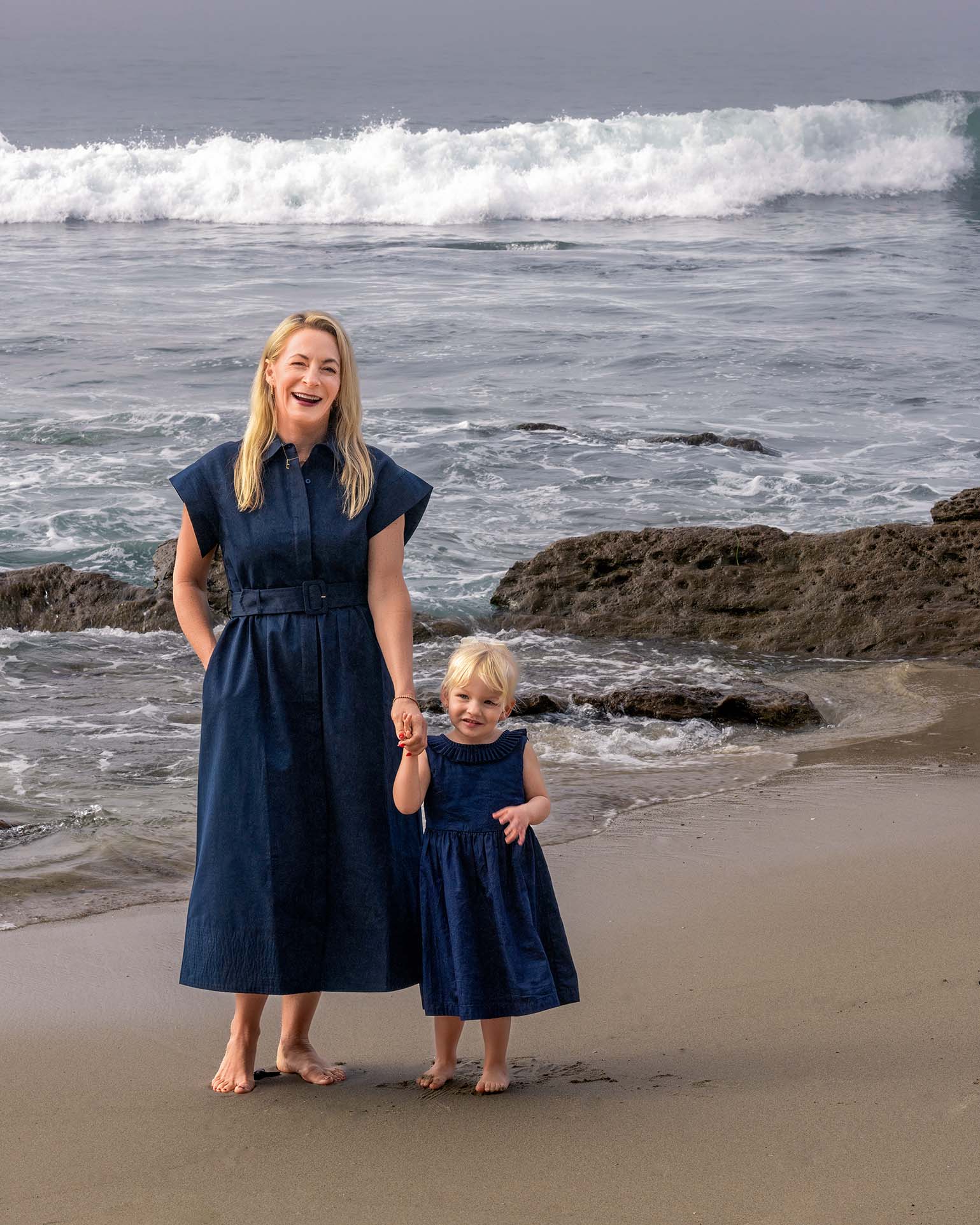 A mom and a young daughter are in blue dresses and standing on the beach.