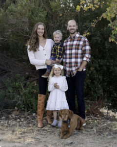 This is a family of two adults and two children and one dog standing in front of trees.