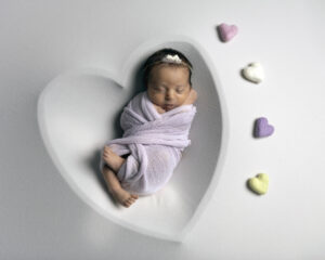 A nwborn baby in Walnut Creek is sleeping in a heart shaped bowl. There are 4 small hearts next to her.