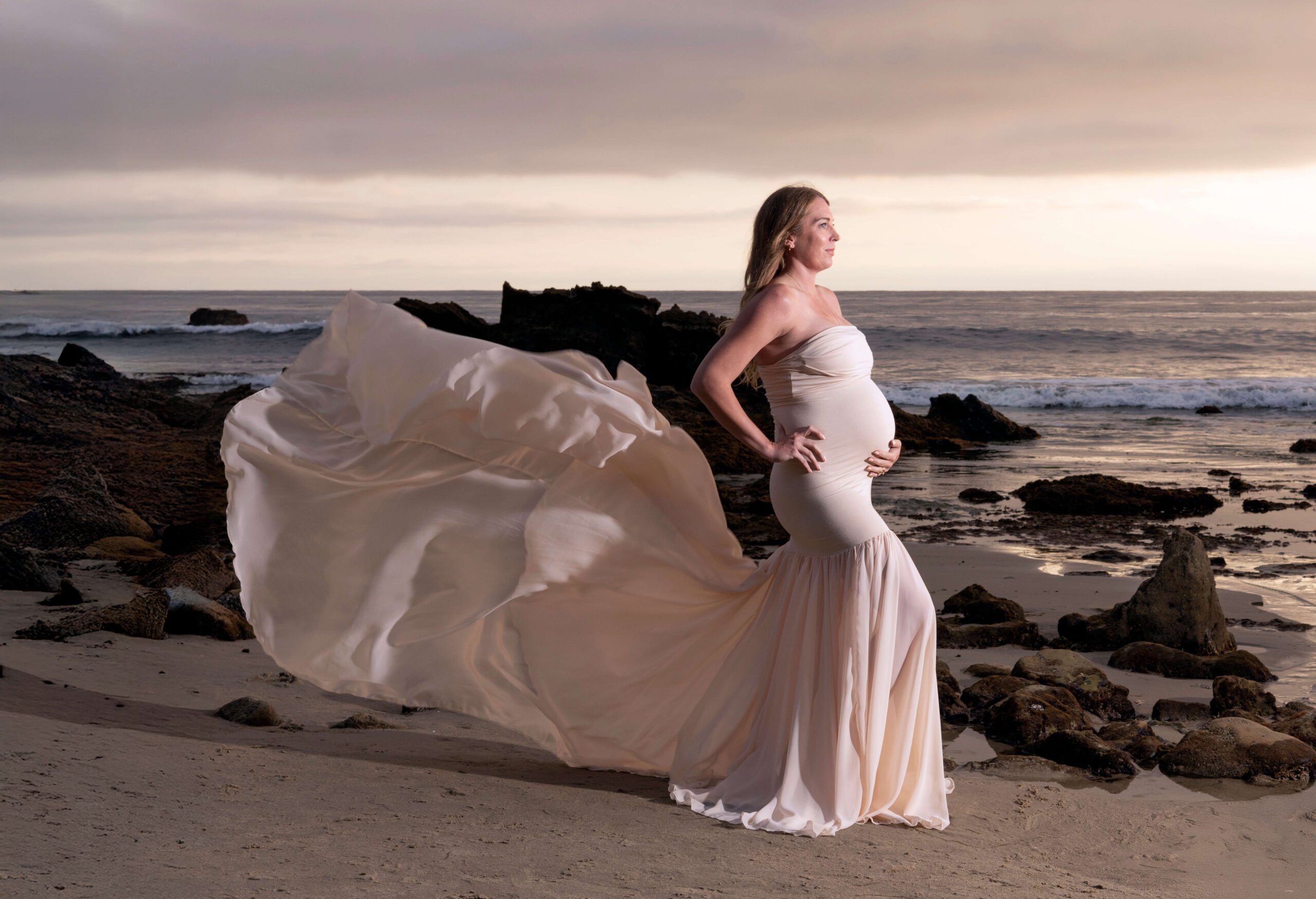 A mother to be stands on a beach at sunset in a cream maternity gown as the train flies in the wind with a hand under her bump after finding doulas in the bay area