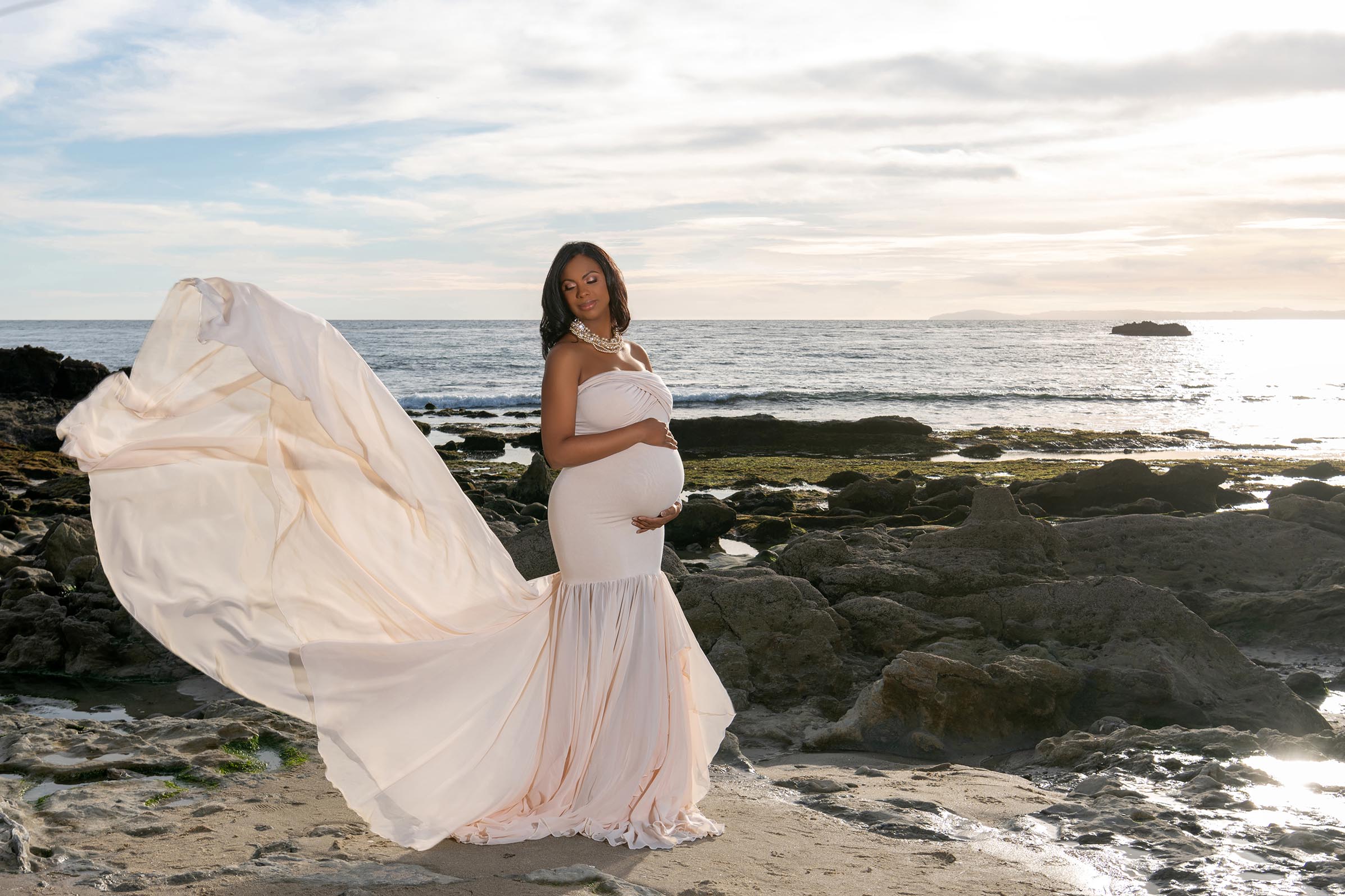A pregnant woman is standing on the beach and her dress is blowing in the ocean breeze.