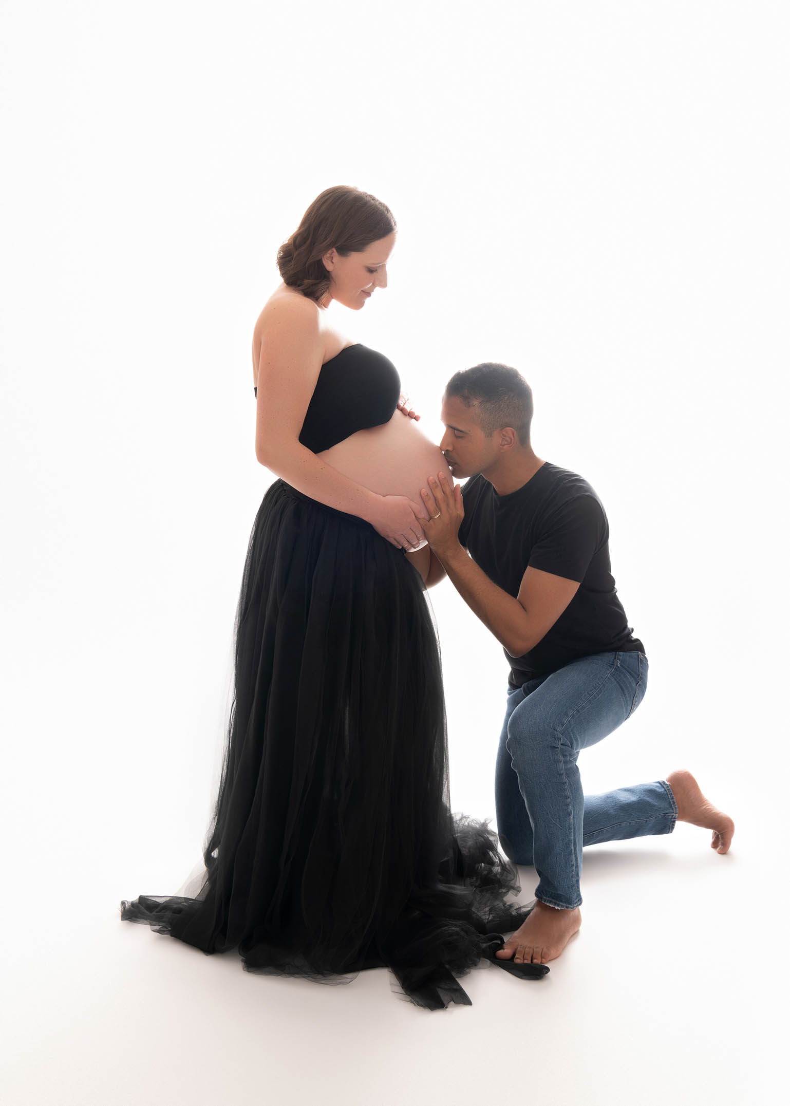 A pregnant woman is wearing a two-piece black gown and her partner is kissing her belly.