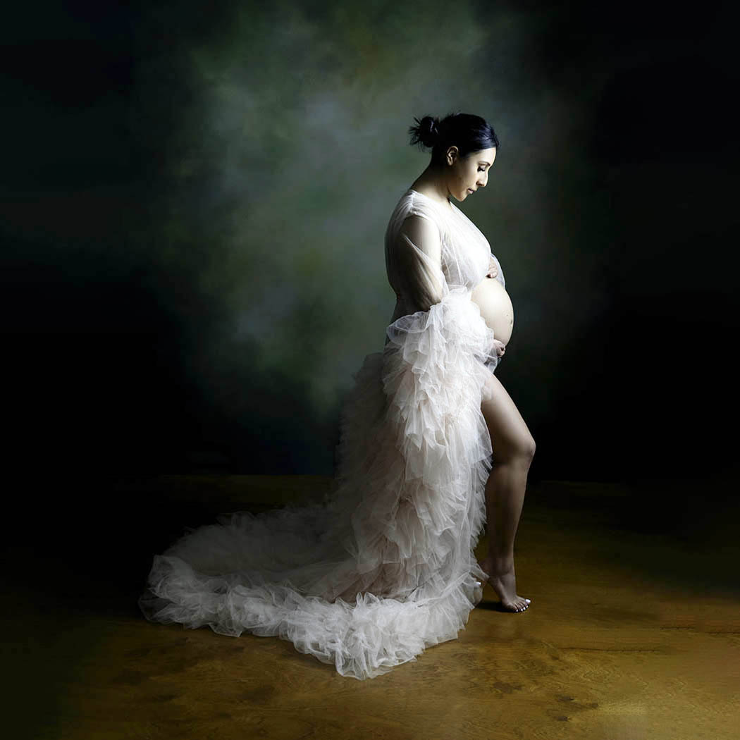 A pregnant woman is wearing a light pink ruffled gown and is looking at her belly.