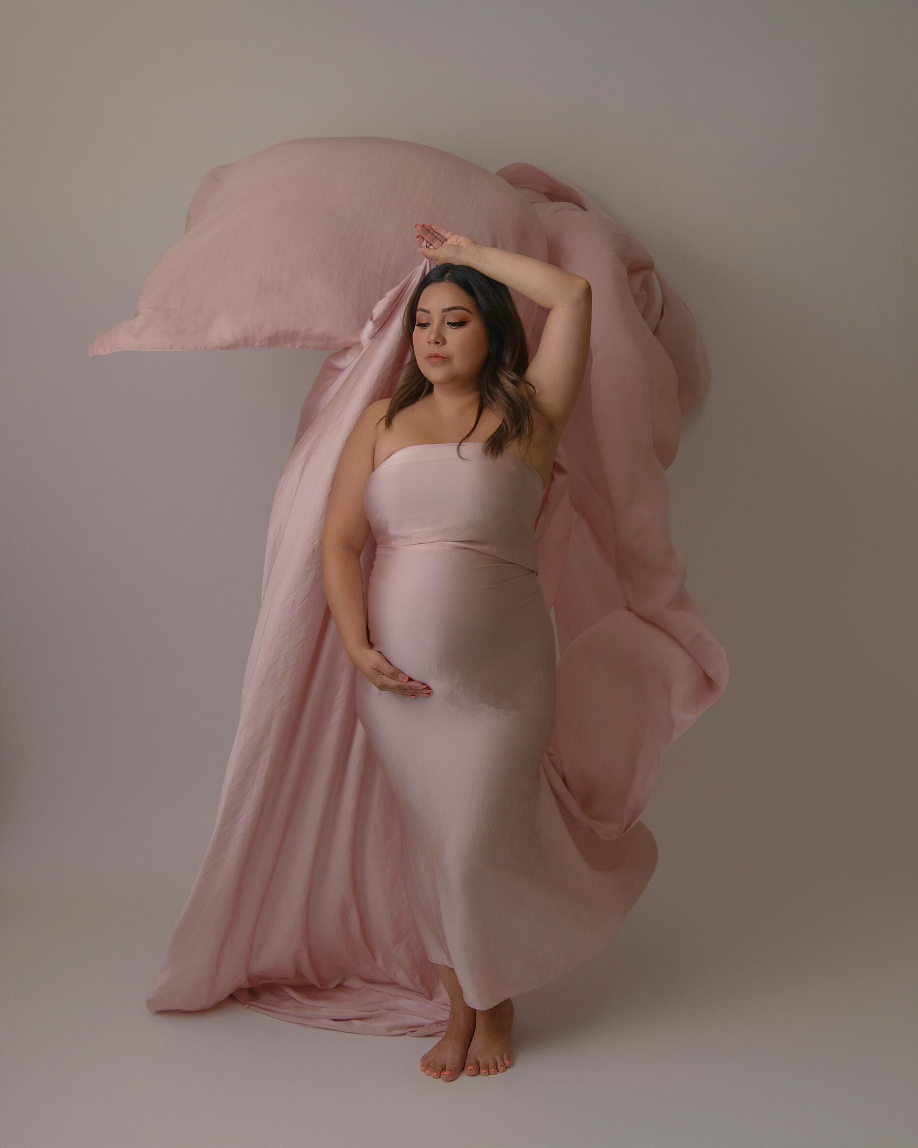 A pregnant woman plays with a long pink silk in a studio with a hand under her bump