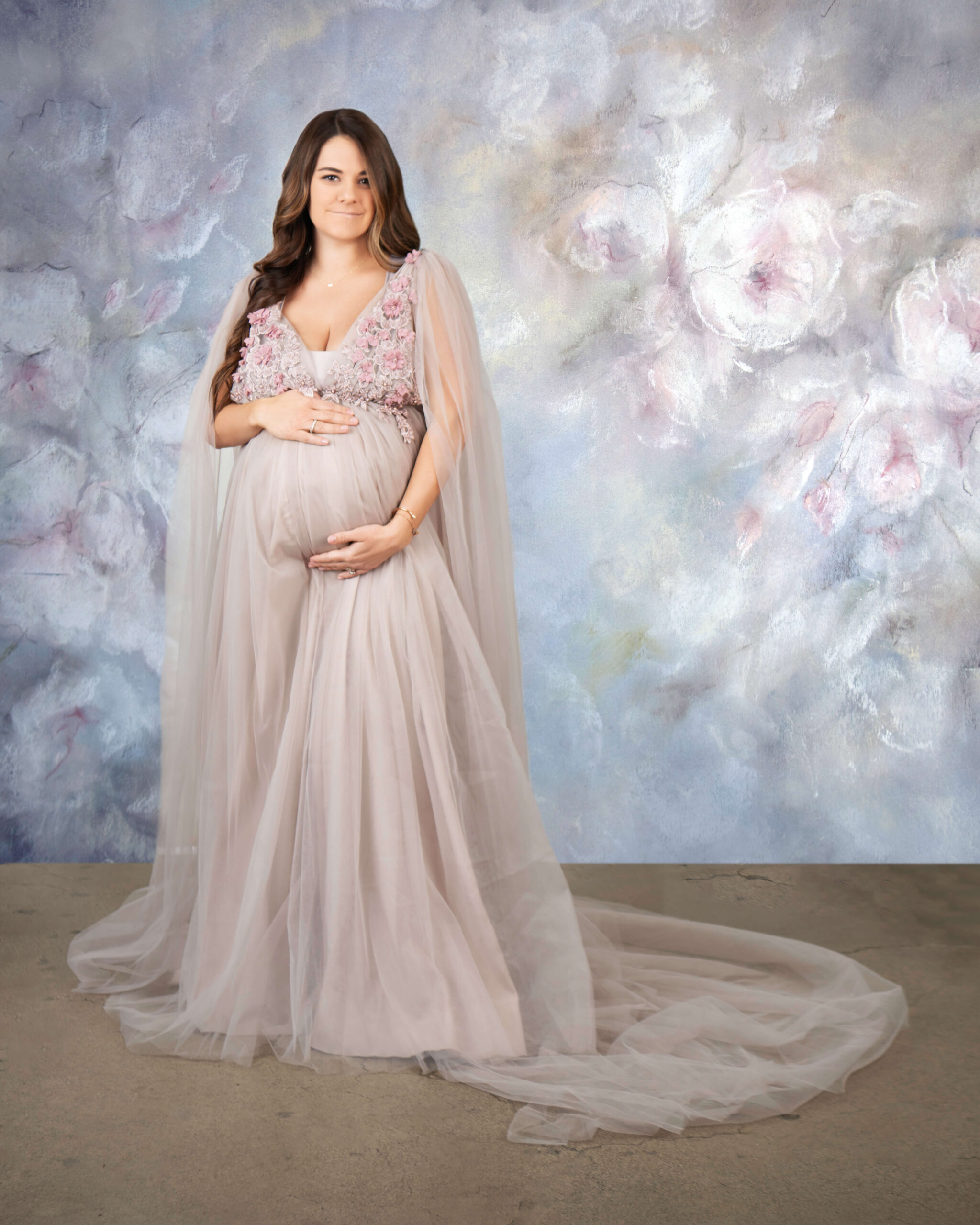 A pregnant mother to be in a long smoky maternity gown stands smiling in a studio with hands on her bump