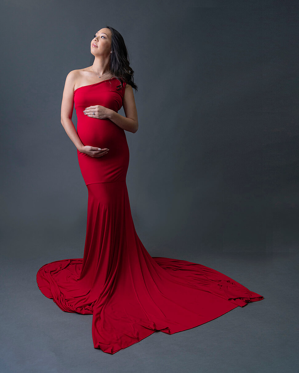A mother to be in a long red maternity gown stands with hands on the bump in a studio