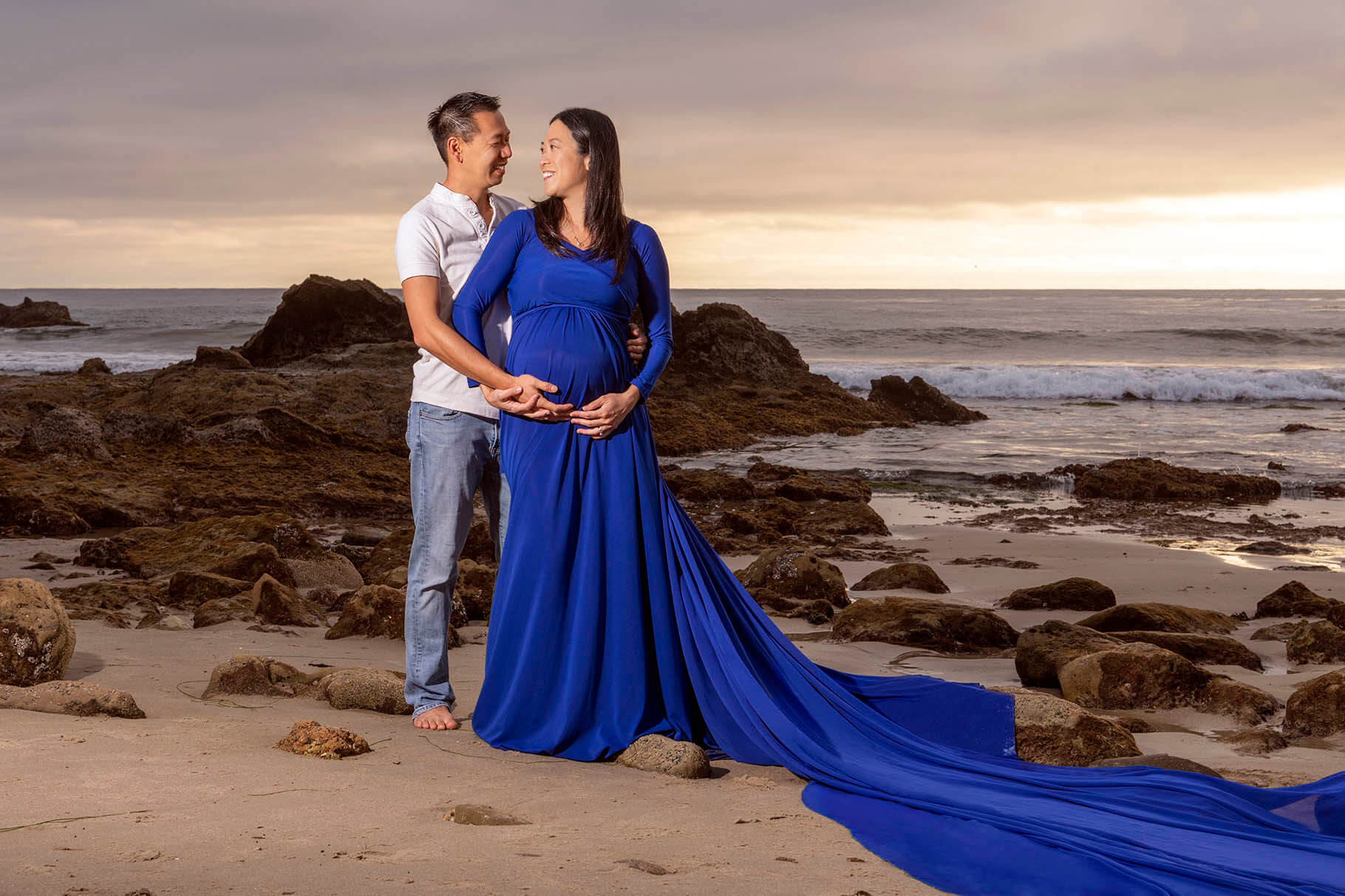 A happy expectant couple stand on a rocky beach at sunset smiling at each other and cuddling