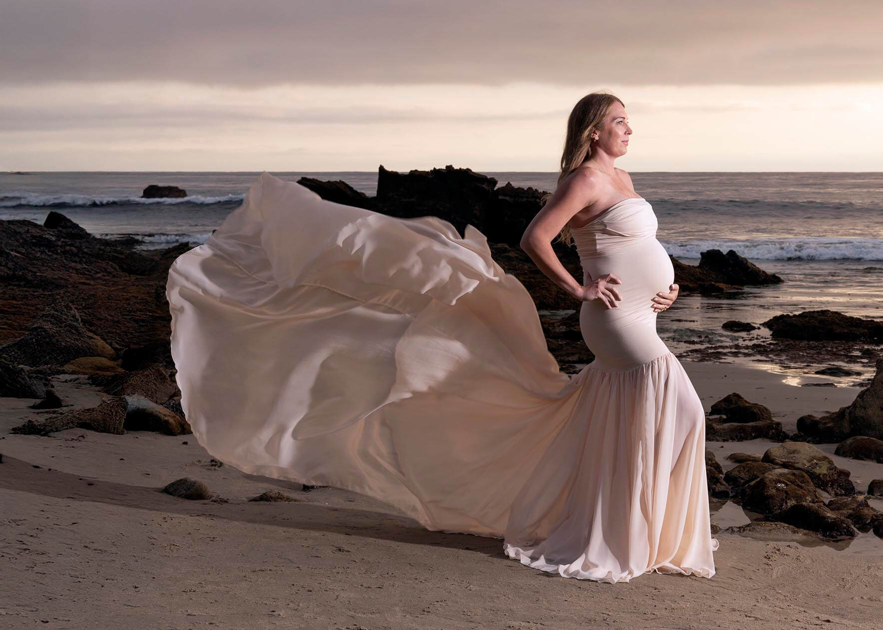 A happy pregnant woman stands in a long flowing maternity gown on a beach at sunset with a hand on her hip after enjoying prenatal yoga in san francisco