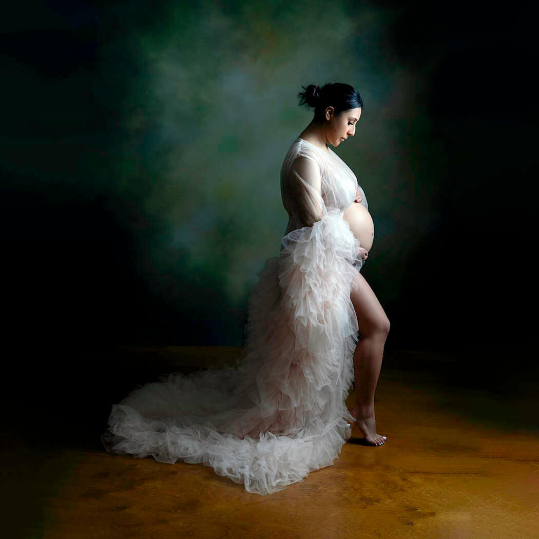 A mother to be in a long tulle maternity gown gazes down at her bump while standing in a studio after some prenatal yoga in san francisco