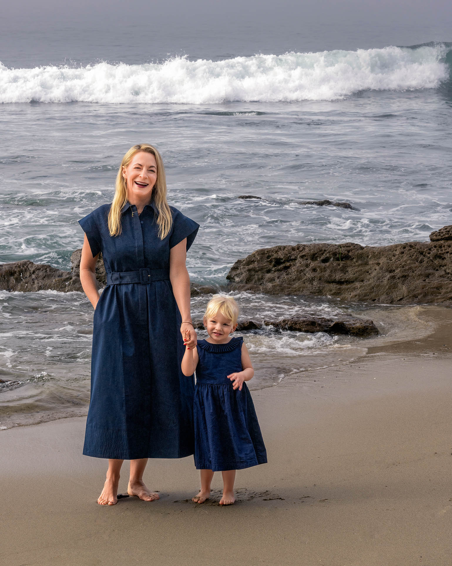 A mom and young daughter are wearing navy blue velvet dresses and are standing on the beach.